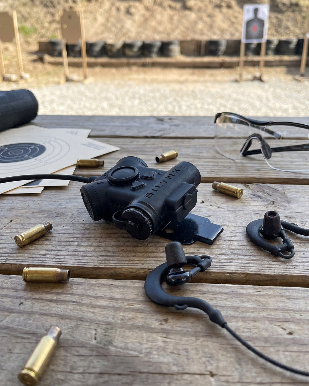 Silynx Clarus II Pro shown on a wooden table at a shooting range with bullet casings, safety glasses, earplugs, and targets in the background.