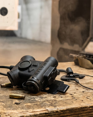 Silynx Clarus II Pro. Resting on a wooden table with bullet casings and a headset nearby in a blurred workshop or shooting range setting.