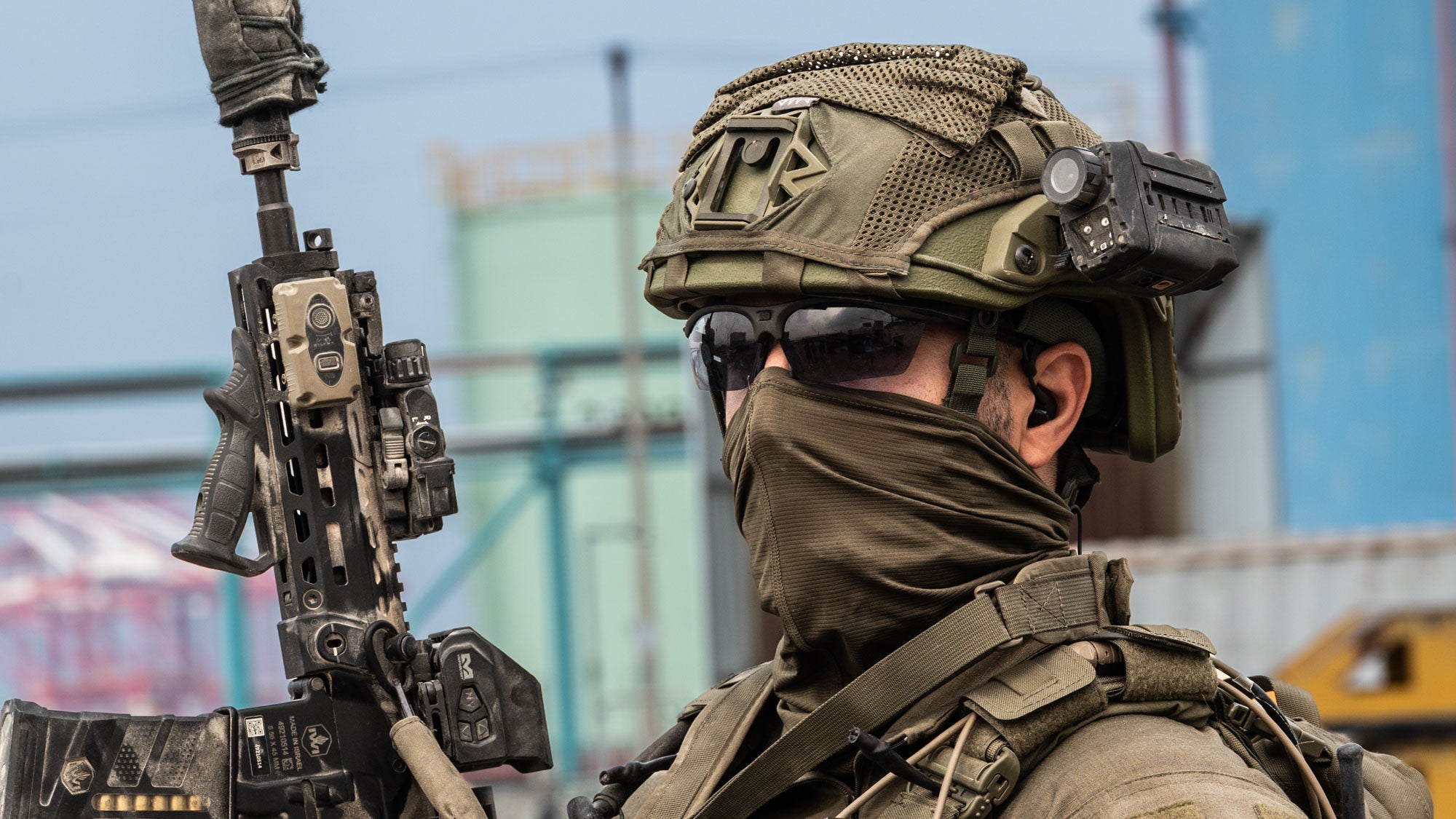 A close-up side profile of a soldier with SIlynx MWPTT and Protego headset with a green tactical helmet with a night vision device, sunglasses, a face covering, and a tactical vest, holding a rifle in an outdoor industrial setting.