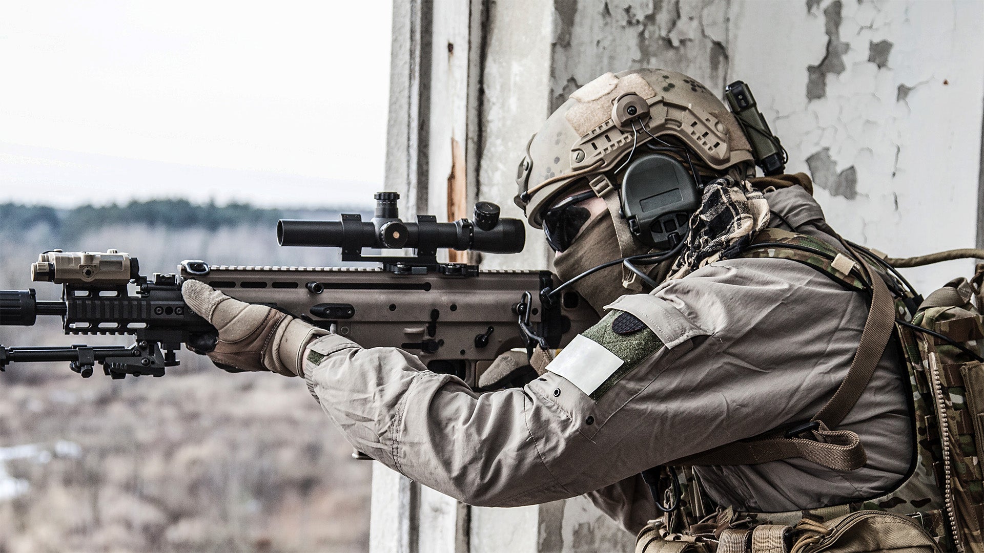 A soldier in full tactical gear, including a helmet, face mask, and rifle with a scope, aiming a rifle from a prone position against a weathered concrete wall.