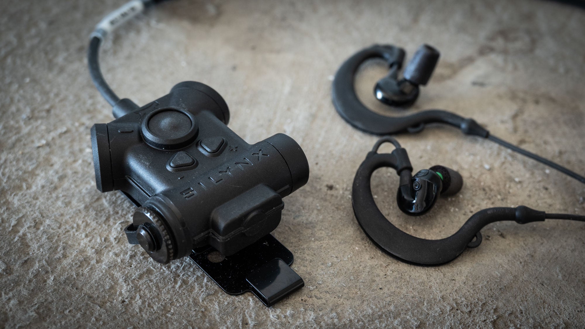 A black Clarus II pro tactical headset shown alongside matching black Protego in-ear earphones on a textured concrete surface.