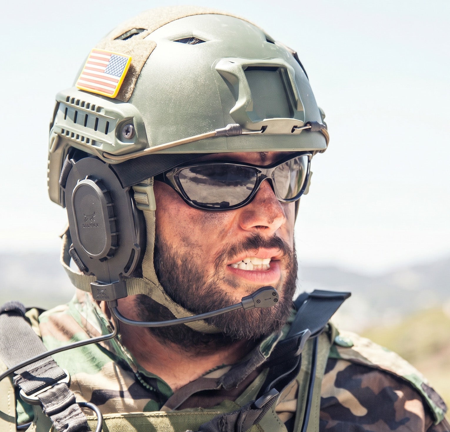 Soldier wearing Silynx HSI Headset on a  helmet with an American flag, sunglasses, against a blurred natural background.