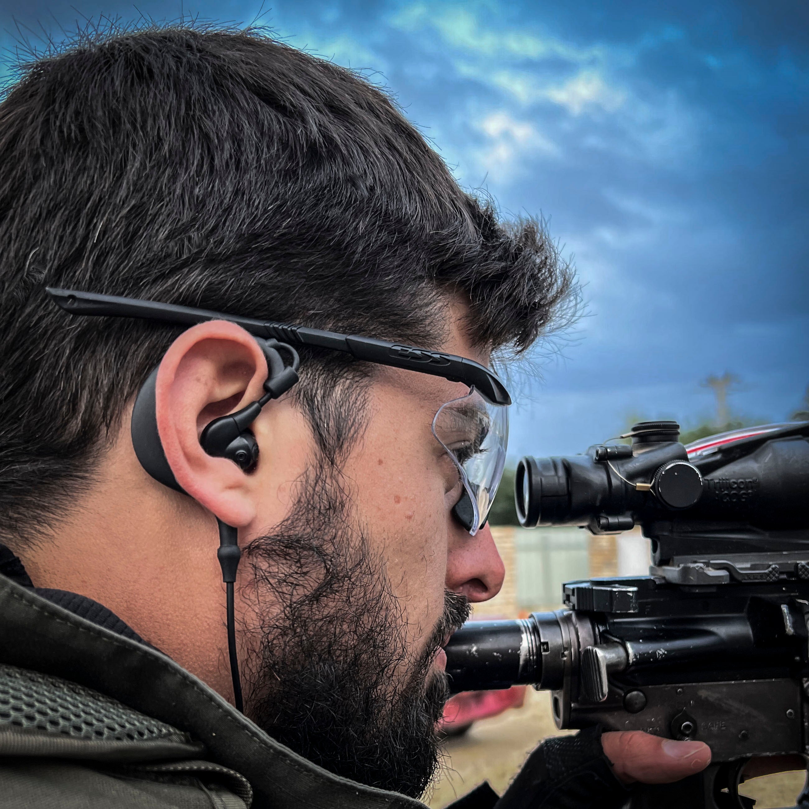 Soldier using Silynx Protego headset with a rifle and a scope and ear protection against a cloudy sky.