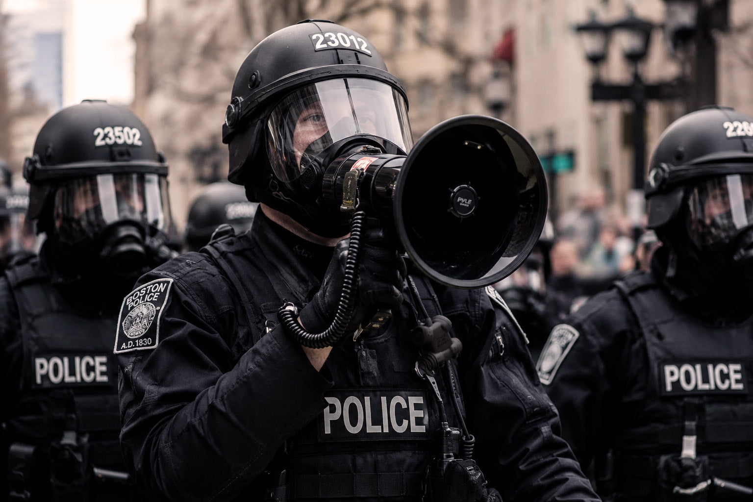 Police officer Silynx Clarus control box and  in full gear with a megaphone, surrounded by other officers in a city setting.