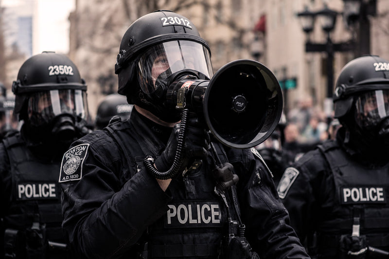 Police officer Silynx Clarus control box and  in full gear with a megaphone, surrounded by other officers in a city setting.