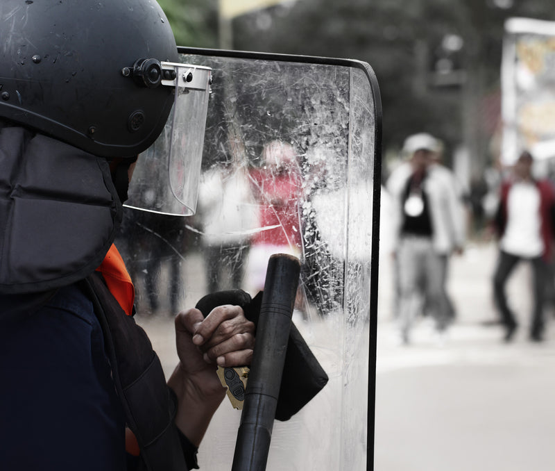 Police man in riot gear holding a shield with Silynx Wireless PTT. Blurred background of people on a street.
