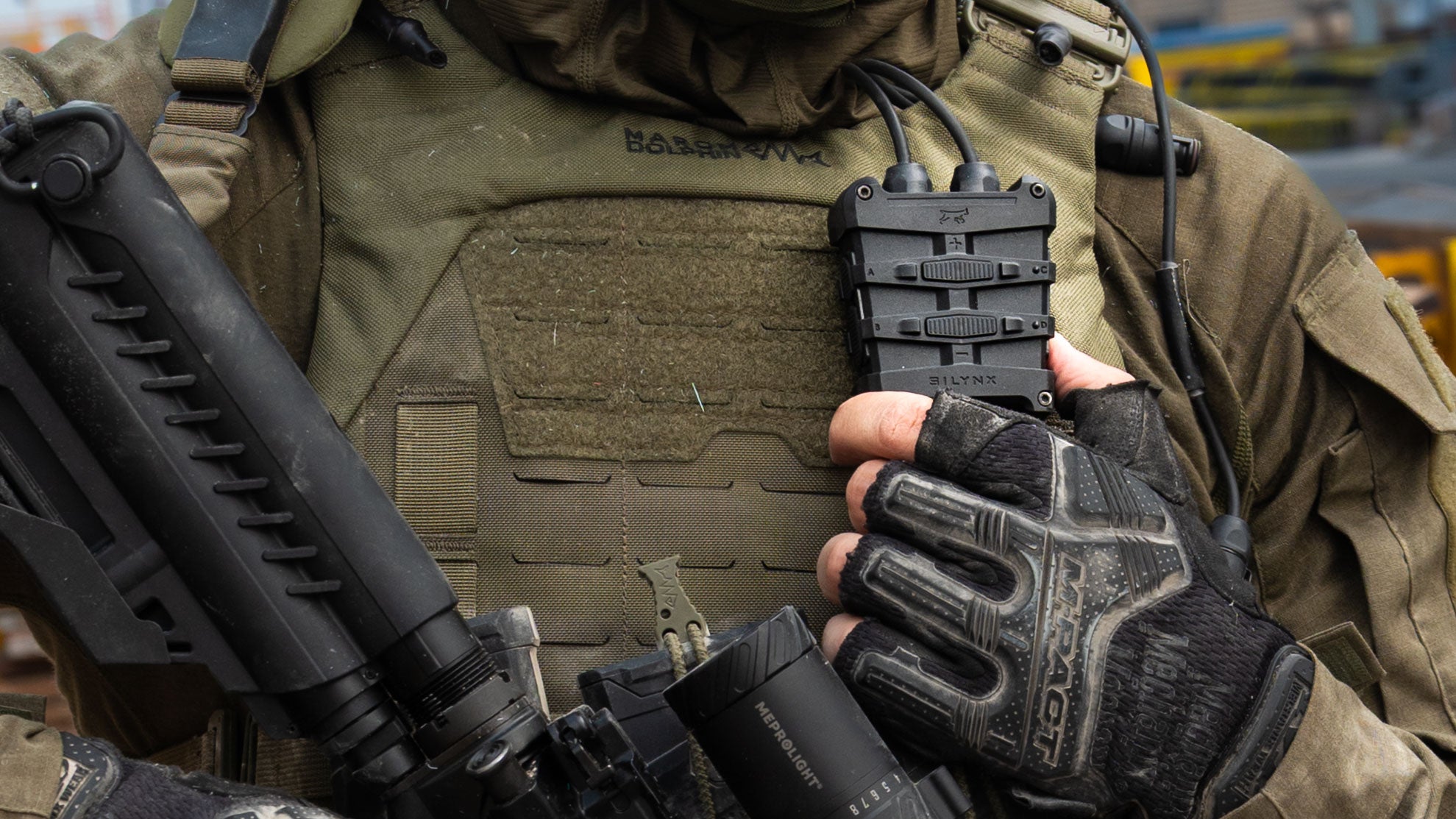 A close-up of a tactical person wearing a black Magellan-branded glove while holding Silynx Fortis control box device attached to a vest.
