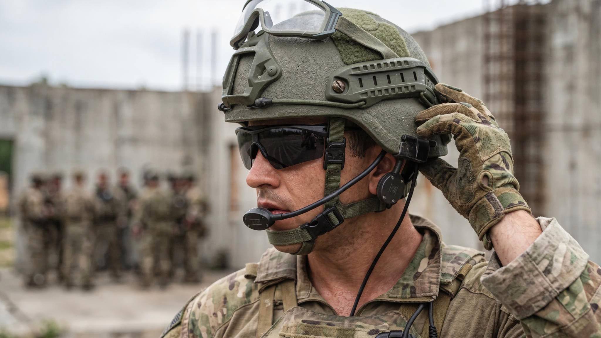 A close-up of a soldier wearing a green tactical helmet, black sunglasses, and  Silynx Eagle Headset headset, adjusting the helmet with one hand. Another soldier is visible in the blurred background.