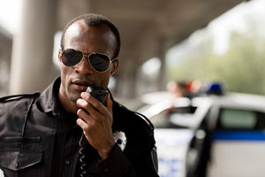 Police man in sunglasses using a speaker mic with a police car in the background